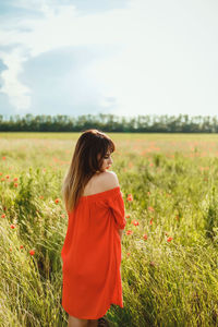 A girl in a red dress above the knee stands in a poppy field during the day against the sky