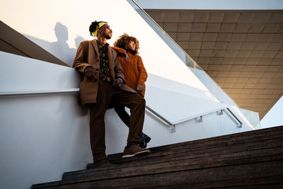 From below of cool african american woman and man in trendy clothes standing together on stairs in sunset light looking away