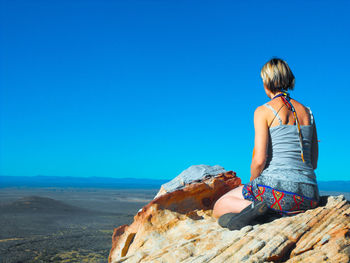 Rear view of woman sitting on rock against clear blue sky