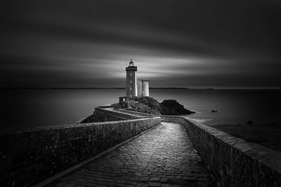 Footpath leading towards lighthouse at sea shore against cloudy sky at dusk