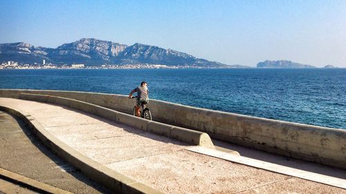 Rear view of man standing on retaining wall by sea against clear sky