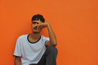 A hand indian young guy looking sideways while sitting outside against orange wall with copy space