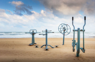 View of fitness equipment on sandy beach against cloudy sky