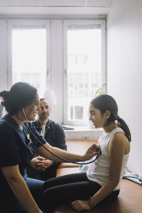 Smiling female nurse listening to heartbeat of patient sitting on bed at hospital