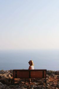 Woman sitting on bench at cliff by sea against clear sky