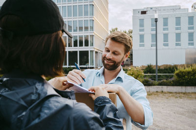 Smiling man signing on document while receiving parcel from delivery person