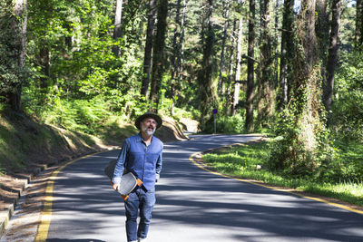 Man with a longboard skateboard in his hands walks along a road in the forest