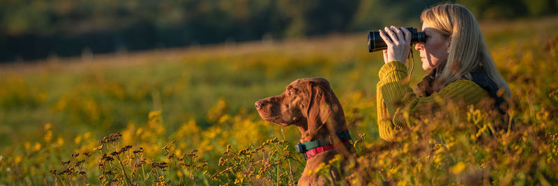 Young female hunter using binoculars for bird spotting with hungarian vizsla dog by her side.