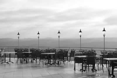 Empty chairs and tables at street against cloudy sky