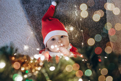 Toddler dressed as santa claus lies under the christmas tree and holds a garland