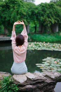 Rear view of woman with arms raised sitting on rock by lake at park