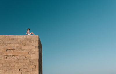 Low angle view of woman sitting on building against clear blue sky