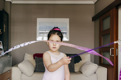 Focused cute little brunette girl in leotard looking at camera while spinning ribbon during rhythmic gymnastic practice training in cozy living room at home