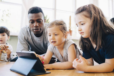 Male teacher looking at girl using digital tablet by friend at table in classroom