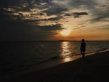 Silhouette man walking on shore at beach against orange sky during sunset