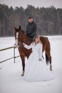 Portrait of smiling groom sitting on horse by bride on snow covered field