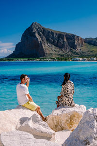 Friends sitting on rock by sea against blue sky