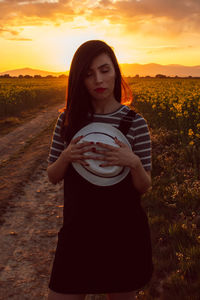 Young woman holding hat while standing on land during sunset