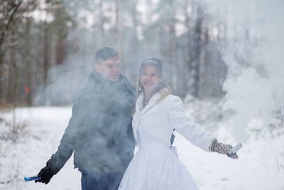 Smiling wedding couple holding distress flares while standing at forest during winter