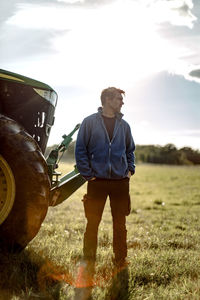 Farmer with hands in pockets standing by tractor on sunny day