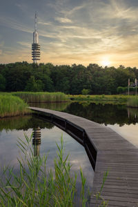 Scenic view of boardwalk crossing lake against sky at sunset with transmission tower in background 