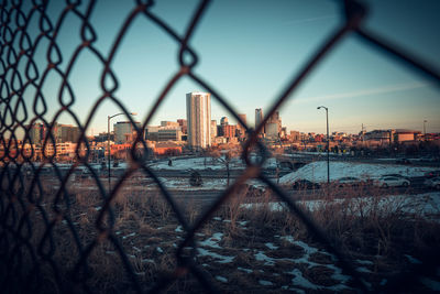 Cityscape seen through chainlink fence against sky during sunset