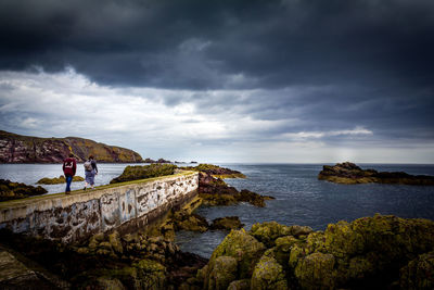 People walking on retaining wall by sea against cloudy sky