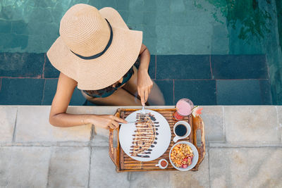 From above of anonymous female tourist in straw hat sitting in pool while cutting delicious crepe with chocolate sauce