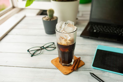 Cold iced coffee in tall glass on bamboo stand against the background of office desk with computer