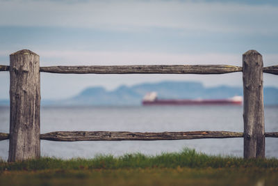 Wooden railing on grass by sea against sky