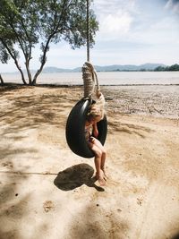 Full length of girl sitting in tire swing at beach against sky