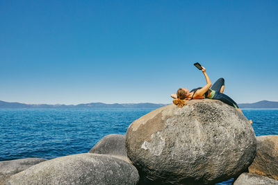 Man on rock by sea against clear blue sky