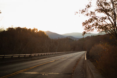 Empty road along trees and mountains against clear sky