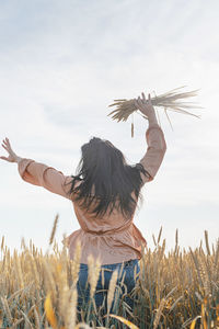 Mid adult woman in beige shirt walking across golden field holding heap of rye lit by sunset light