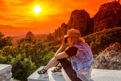 Woman photographing on retaining wall against mountains during sunset