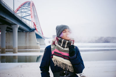 Young woman smoking while standing by bridge against sky during winter