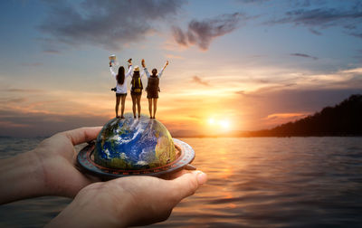 Digital composite image of hands holding planet earth with friends standing while looking at sunset over sea