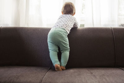 Rear view of baby girl looking through window while standing on sofa at home