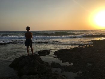 Rear view of man standing on beach against sky during sunset