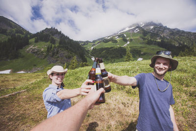 Happy friends toasting beer bottles on field against mountains and cloudy sky