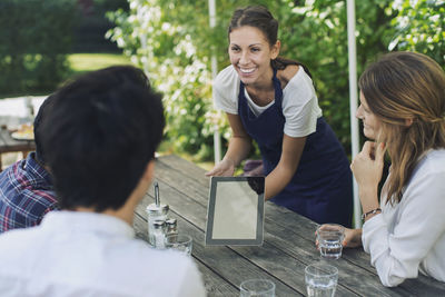 Happy waitress showing menu through digital tablet to people at outdoor restaurant