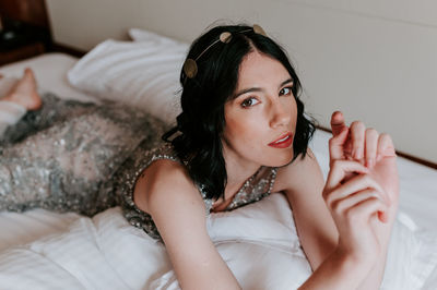 Calm female in sparkling elegant dress resting on comfortable bed with pillows while looking at camera