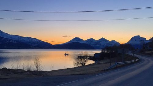 Empty road by river leading towards snowcapped mountains at sunset