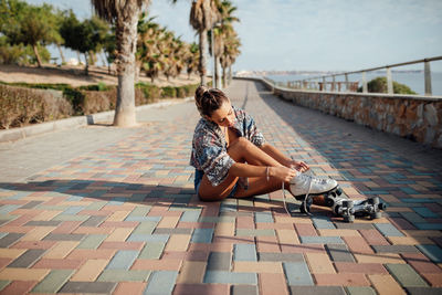 Full body side view of sportive female tying laces on roller skates while sitting on paved walkway before training in city