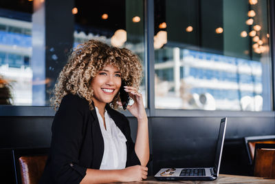 Smiling hispanic afro business woman in cafe working on laptop and mobile phone. tech and lifestyle