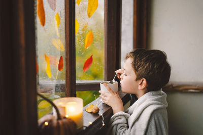 Boy wrapped in a blanket drink hot tea and looks out the open window at the wonderful autumn nature