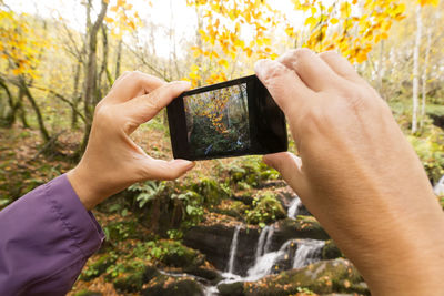 Midsection of person photographing stream in forest during autumn