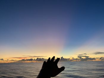 Silhouette person hand by sea against sky during sunset