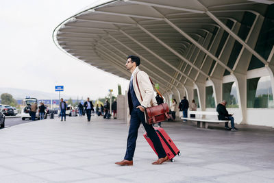 Young man with dark hair in trendy clothes with suitcase and shopping bags walking near modern airport in sunny day