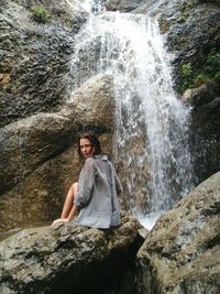 Portrait of young woman sitting on rock against waterfall